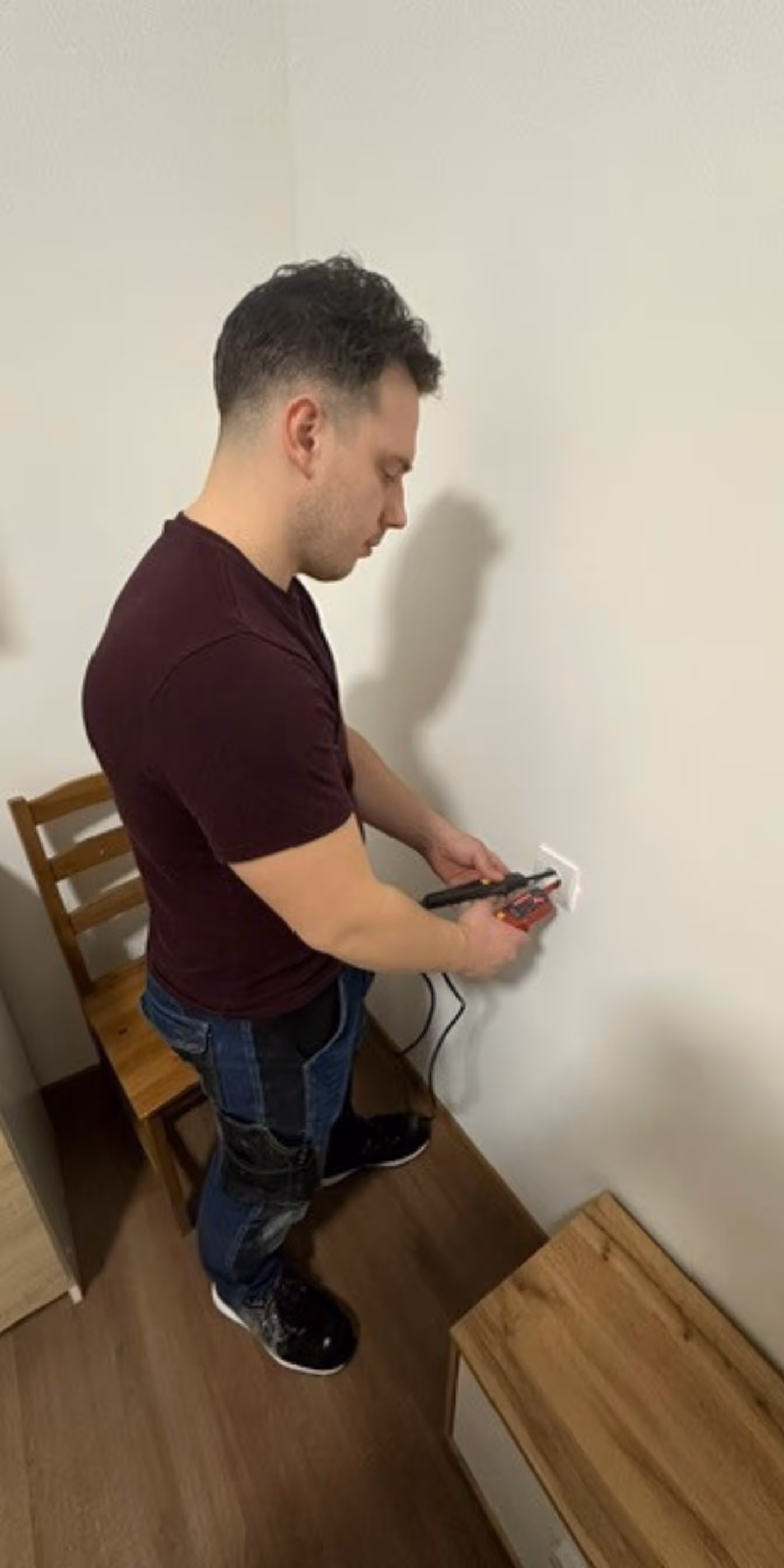 Man in dark shirt and jeans using a voltage tester on a wall electrical outlet in a room with wooden furniture.