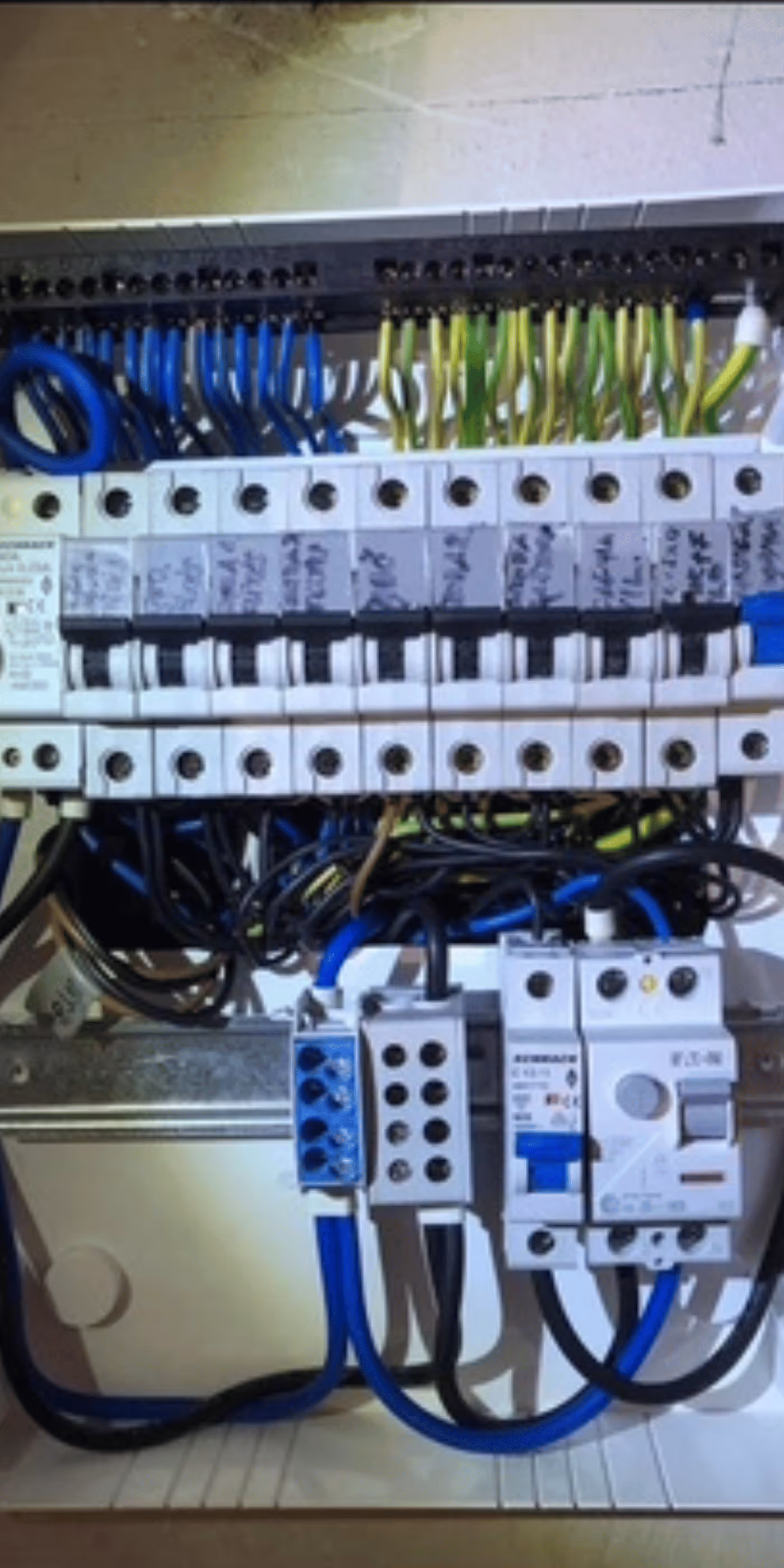 Electrical panel with circuit breakers connected by blue, green, yellow, and black wires inside a white enclosure.