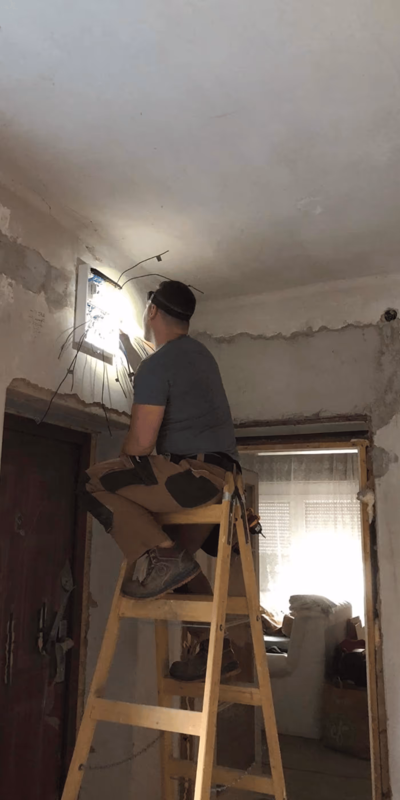 Man sitting on a wooden ladder fixing electrical wiring in a partially renovated room with exposed walls.