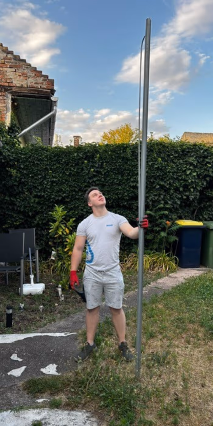 Man wearing red gloves holding and looking up at a tall metal pole in a garden with green bushes and recycling bins behind.